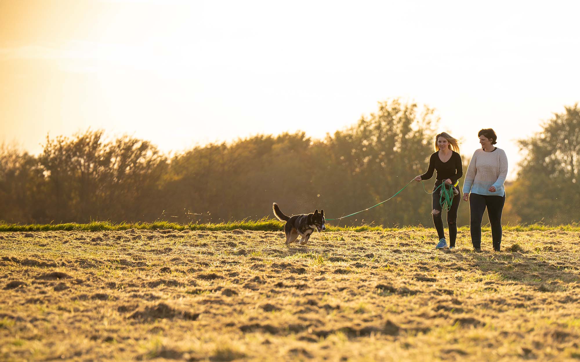 Eva Hoffmann mit einer Kundin und ihrem Hund an der Leine bei Sonnenuntergang auf einem Feld